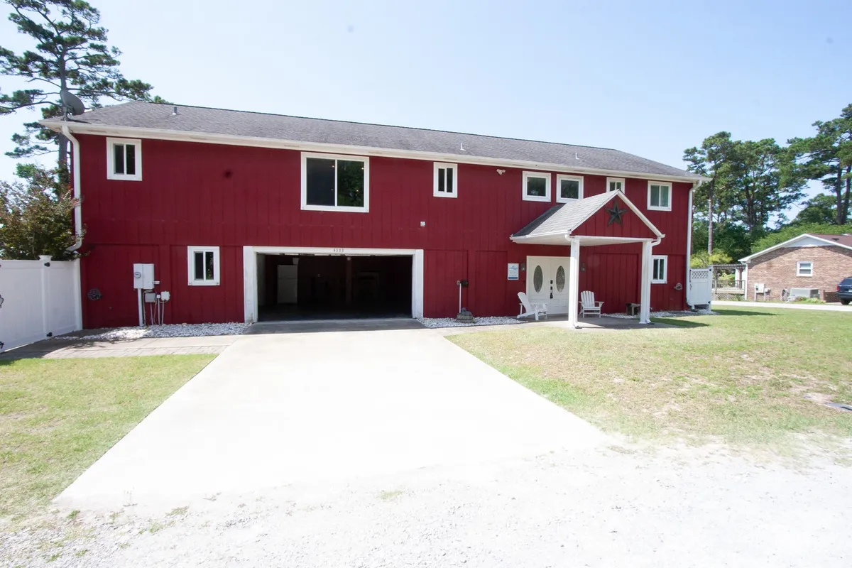 The exterior shows the red house with white trim and a welcoming porch. The open garage space is visible, demonstrating both the ease of parking and the potential for storing adventure gear. The driveway and parking area are in view, indicating space for trailers, boats, or extra vehicles.