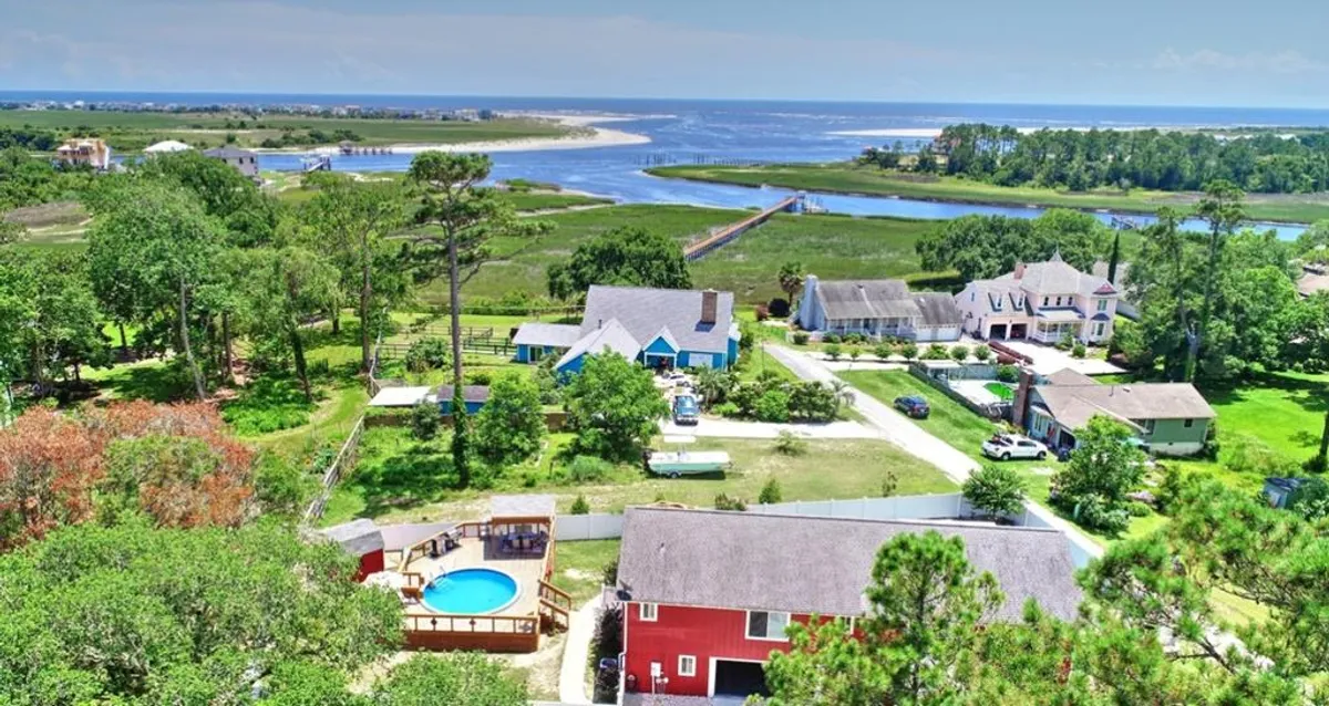 A beautiful exterior view featuring a blue and red house, an above-ground pool surrounded by a sun-soaked deck, and a scenic river landscape. Groups are seen swimming, barbecuing, and relaxing, capturing the essence of outdoor fun and leisure.