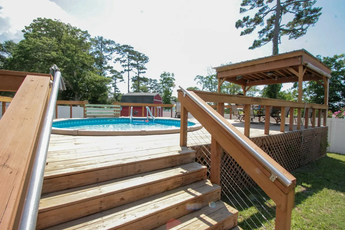 The image showcases the inviting backyard of The Beach Barn, featuring a sparkling swimming pool surrounded by a spacious deck with lounge chairs, lush greenery, and a charming gazebo perfect for gatherings or relaxing out of the sun.