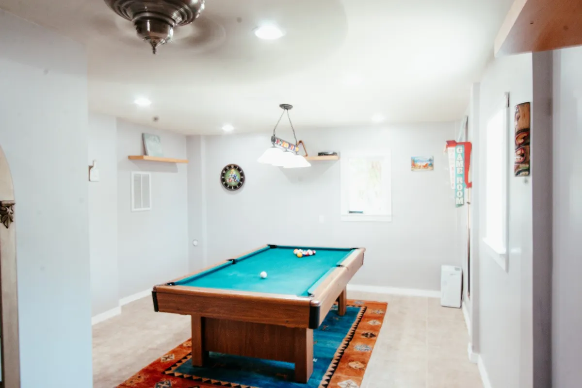 A playful, inviting game room featuring a pool table, dartboard, and colorful wall art. The space is well-lit with a ceiling fan overhead and comfortable seating, perfect for group entertainment and friendly competition.