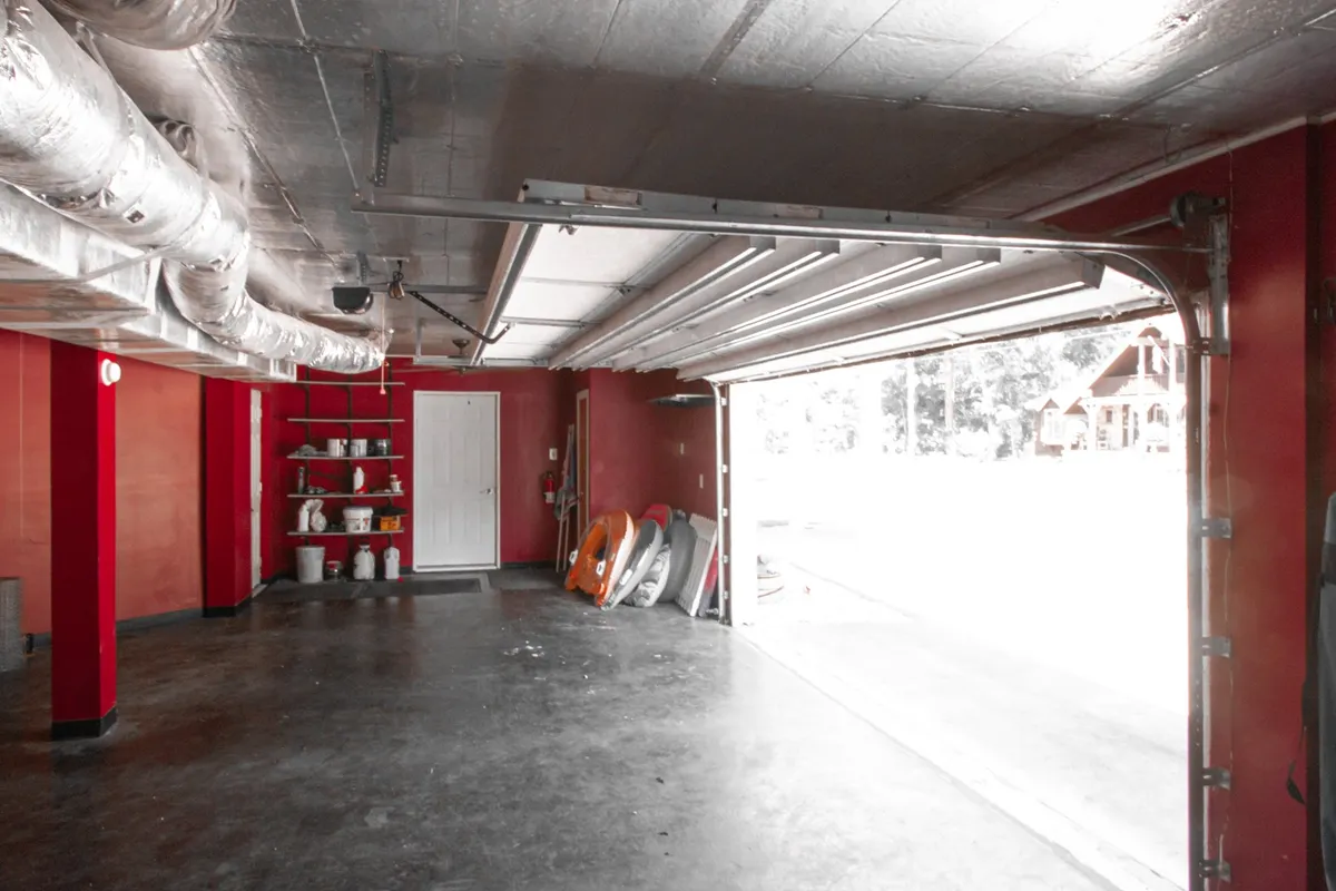 An interior shot of the garage workshop with a large door opening, painted red walls, and shelving units storing tools and adventure equipment. The practical, industrial mood highlights the space's functionality for organizing outdoor gear, such as fishing tackle, paddleboards, or bikes.