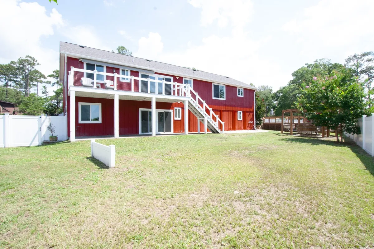 The inviting red exterior of the house, a staircase leading to an upper deck, a lush green lawn, and surrounding trees form the backdrop for family barbecues and games in the fenced yard. The setting is welcoming and relaxed, ideal for both group gatherings and quiet moments.