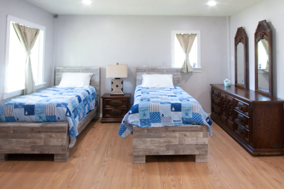 This image shows a tranquil bedroom with two twin beds, a nightstand with a lamp, and a large dresser with mirrors. The room is softly lit by natural light, emphasizing its calm and restful atmosphere.